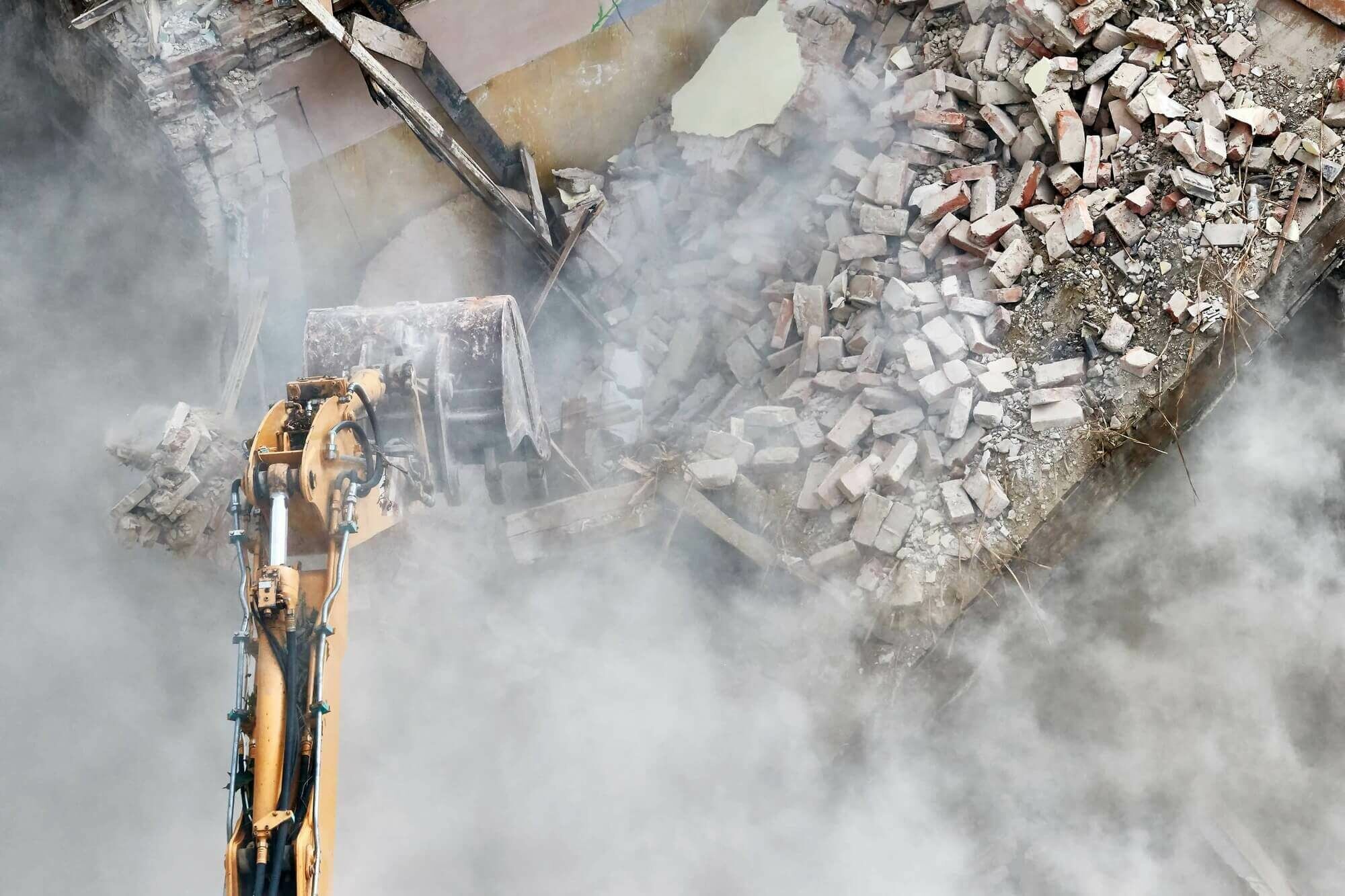 Unsere Leistungen Gebäudeabbruch mit einem Bagger in einer Staubwolke